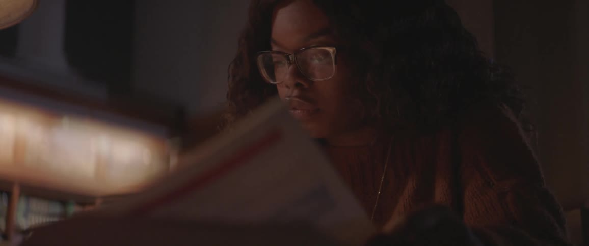 A young black woman wearing glasses is studying a page from a book while she writes down some notes on a piece of paper