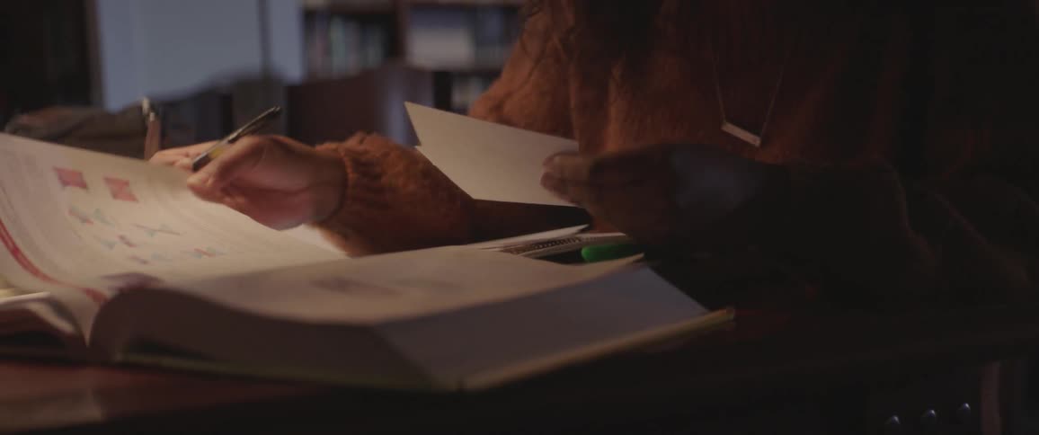 Female turning the pages of a text book while holding note cards in one hand and a pen in the other