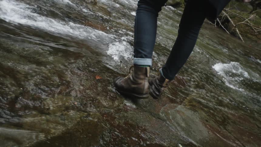 A woman in boots walking into and standing in a shallow stream