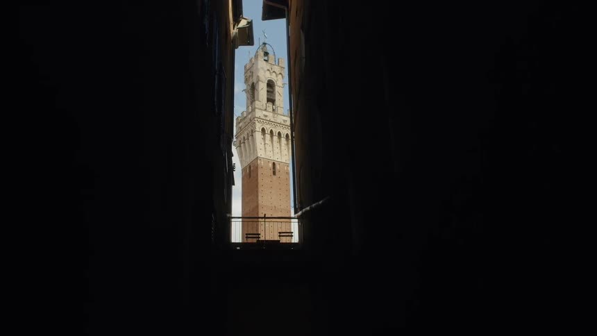A clock tower outdoors is viewed from a tall narrow window in a dark room