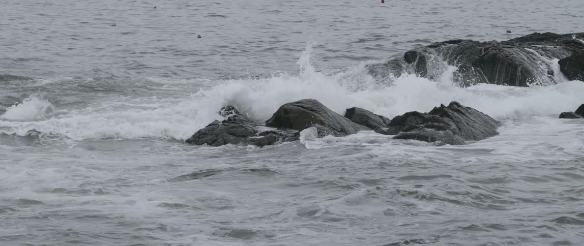 Ocean waves crash forcefully against a group of rocks producing foamy waters on an overcast day