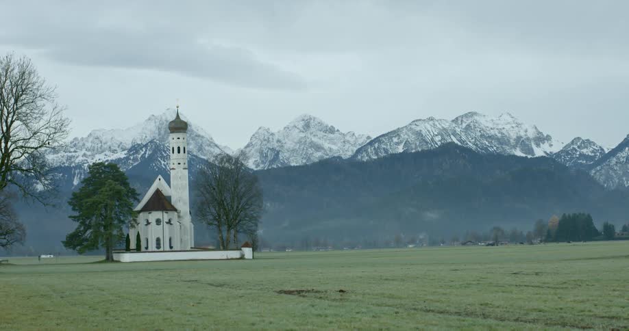 A small russian church sits in a field with snow capped mountains in the background