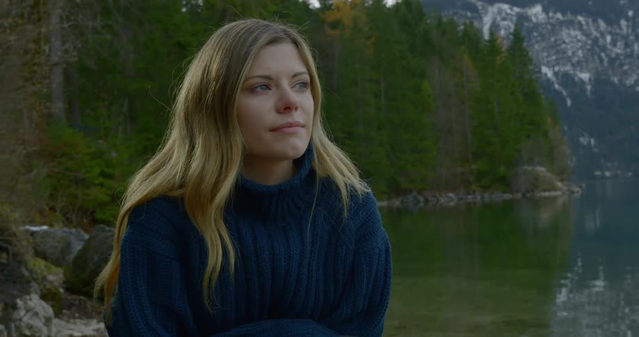 A female with long blonde hair is sitting near the edge of the water with a forest and mountains in the background