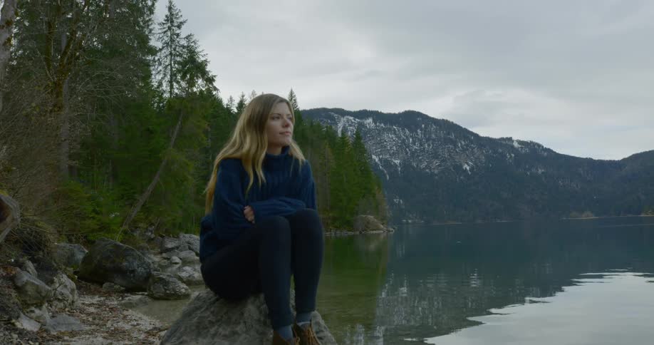 Girl contemplatively thinking while sitting on top of a rock near the lake