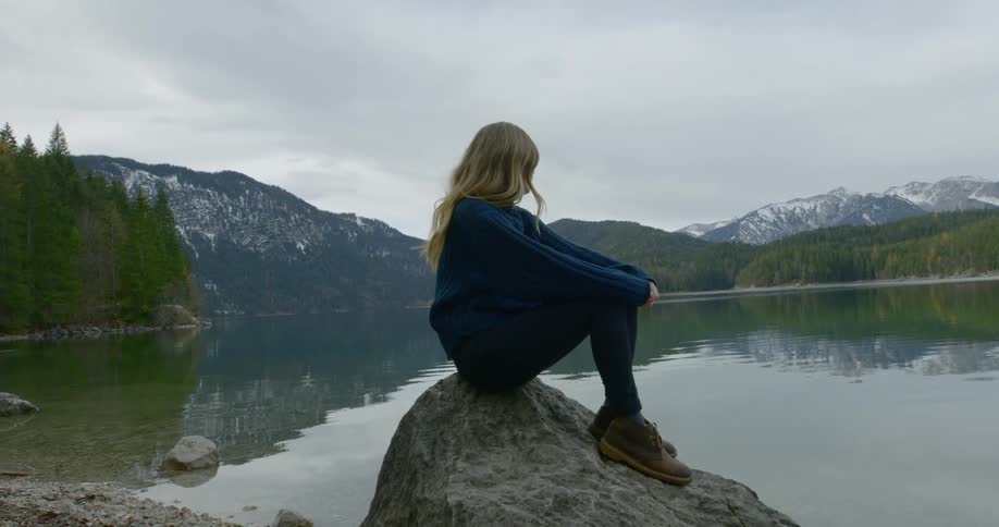 A white female with long blonde hair is sitting on a rock next to a reflective lake with mountains and forest in the distance
