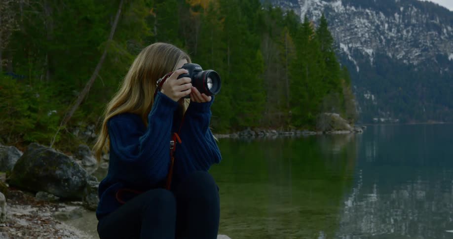 A woman sits by a river and takes a photo with a camera