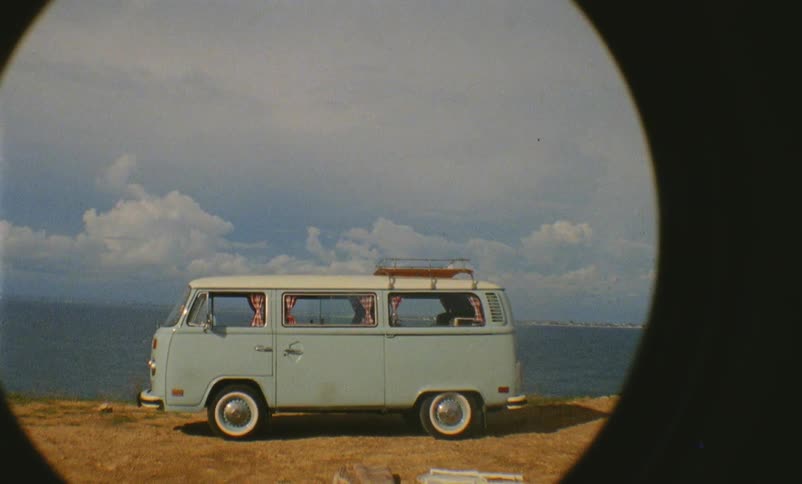 An older model of volkswagen bus is parked next along the shore next to the ocean on a sunny day
