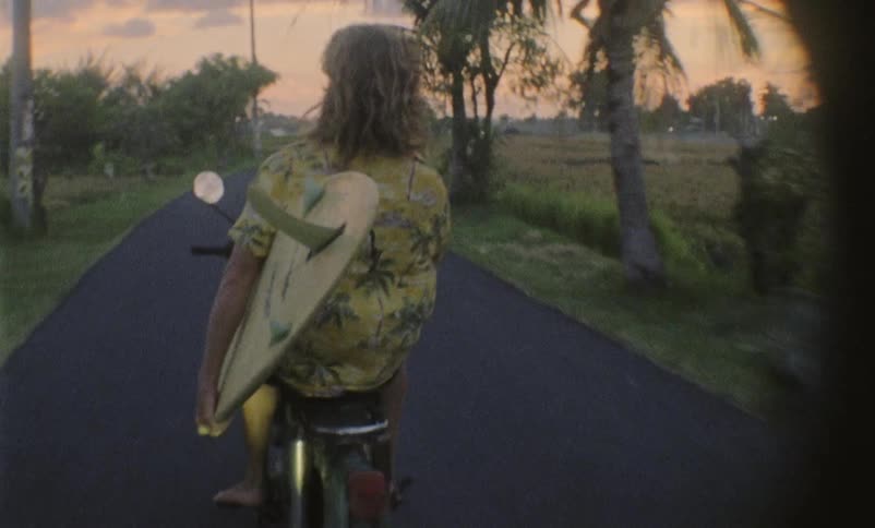 A man rides down the street on a moped carrying a surf board