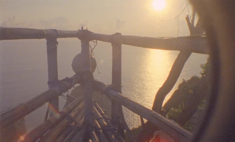 The outline of a camera lens focuses on the ocean below from a rickety wood balcony