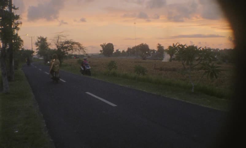 A man carrying a surf board rides by on a motorcycle at sunset on a rural road