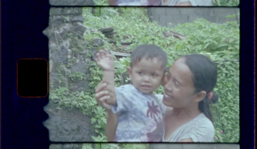 A woman holds a small boy and makes him wave towards a video camera