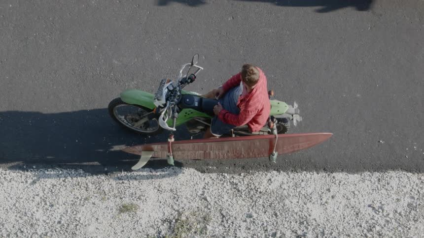A man sits on a green motorcycle with a surfboard strapped to it