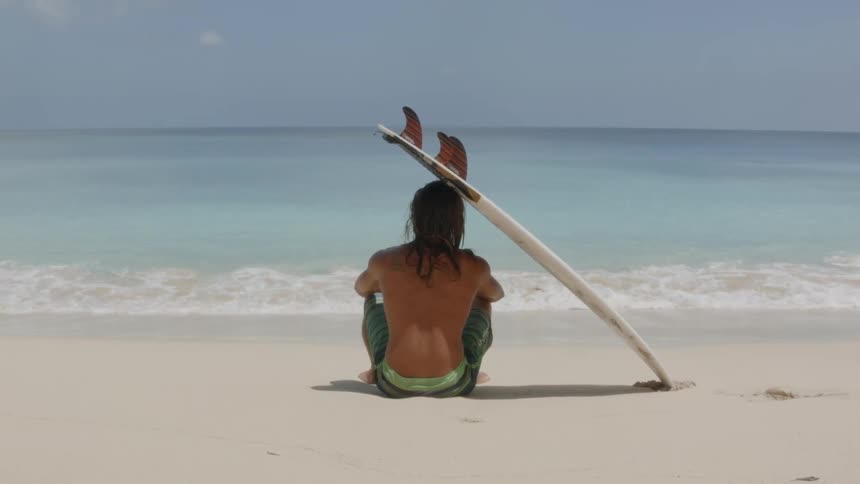 A young man sits on the beach with a surf board resting on his head
