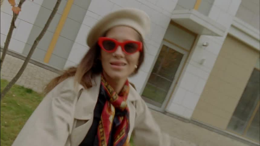 A woman in a long coat dances around in front of a building while wearing a pair of red sunglasses and a beret