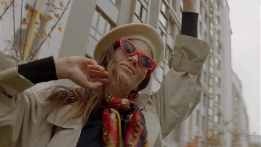 A woman with hat and shades in front of a building frolicking