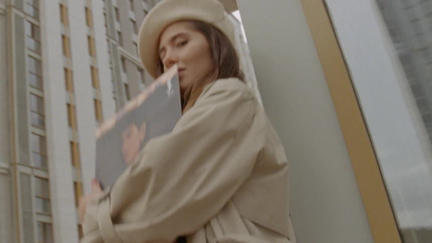 A woman in stylish clothing leaning against the corner of a building holding a vinyl record case