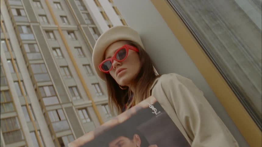 A model holds a poster while leaning on the corner of a building in a city