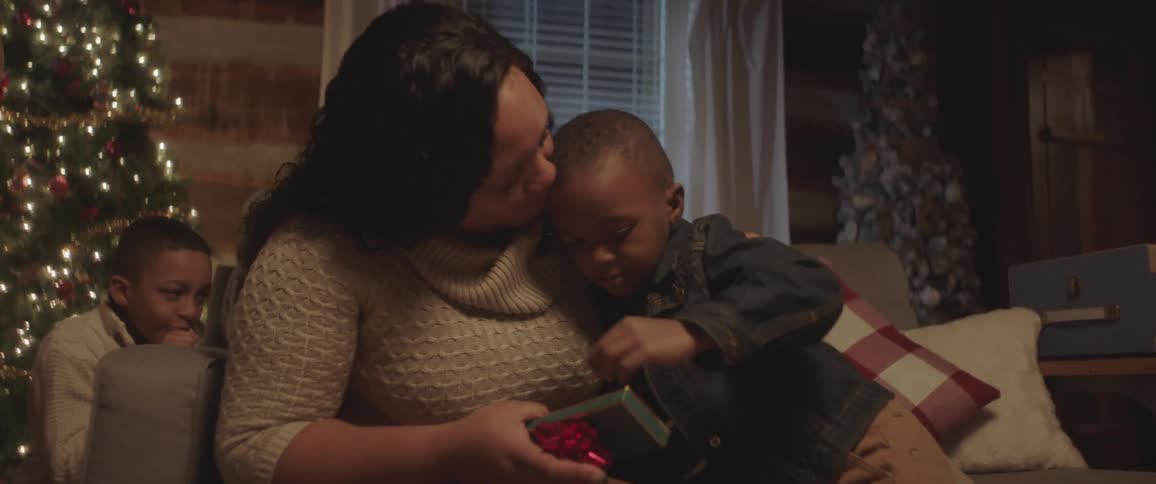 A young boy is given a hug and a kiss from his mother has she opens a christmas gift with her family in front of their decorated tree