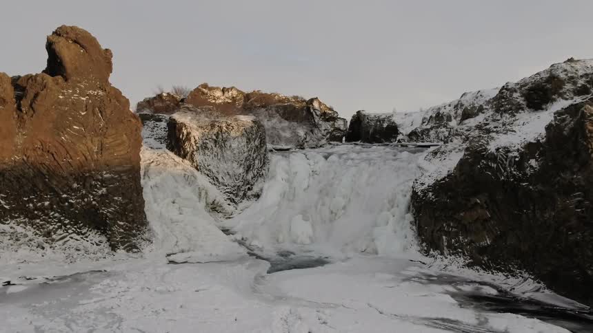 A waterfall partially frozen to ice surrounded by hills and mountains
