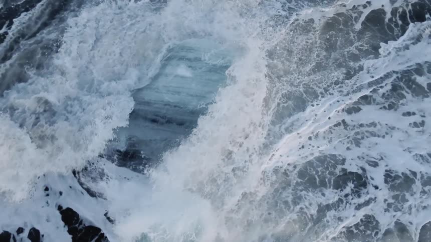 Forceful and large waves crash violently against a large piece of ice that is atop a rock formation that is jutting from the ocean surface