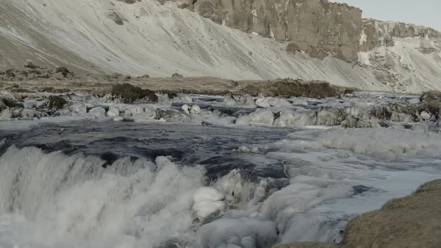 A river runs through the base of a deep canyon in which the water is rushing over ice covered rocks
