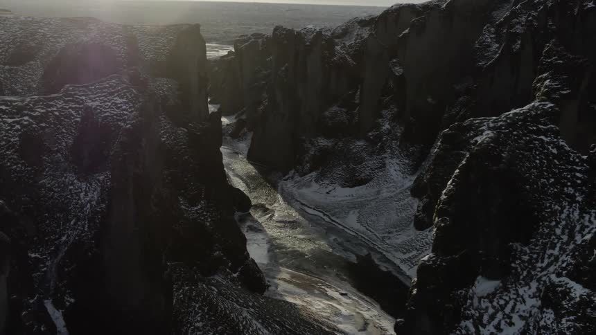 A tall and jagged range of dark mountains are covered in light snow as a river flows in a canyon below
