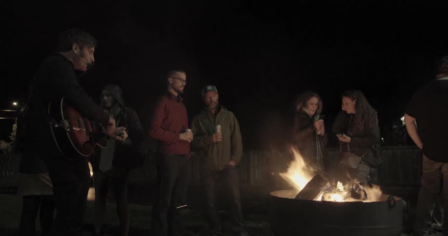 A man plays guitar and sings as people chat around a fire