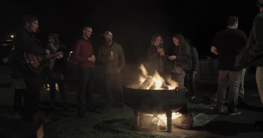 People chat around a bonfire as a man plays guitar and sings
