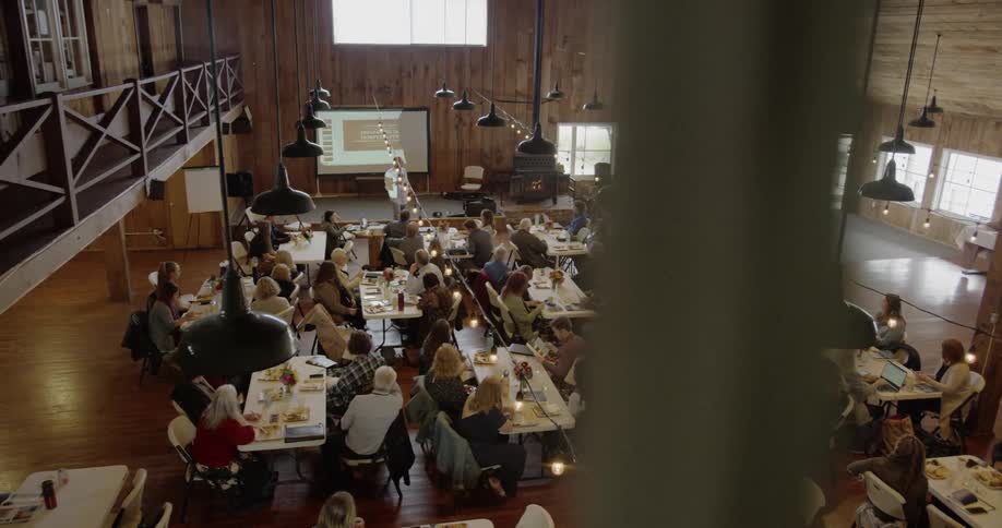 A sizable group of people are gathered at tables indoors while a person stands on a small stage giving a presentation in front of a slide projector
