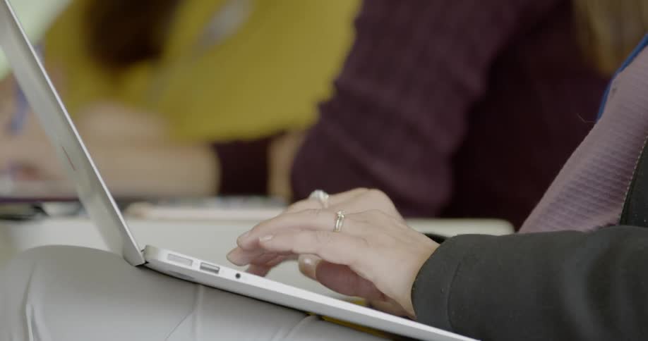 A woman types on a laptop that is sitting on her lap in a classroom
