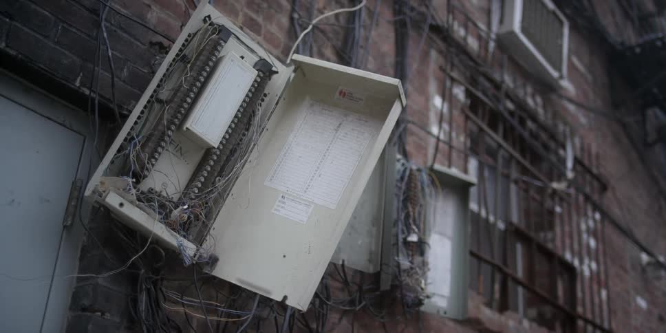 A control box is open and damaged on the side of a building