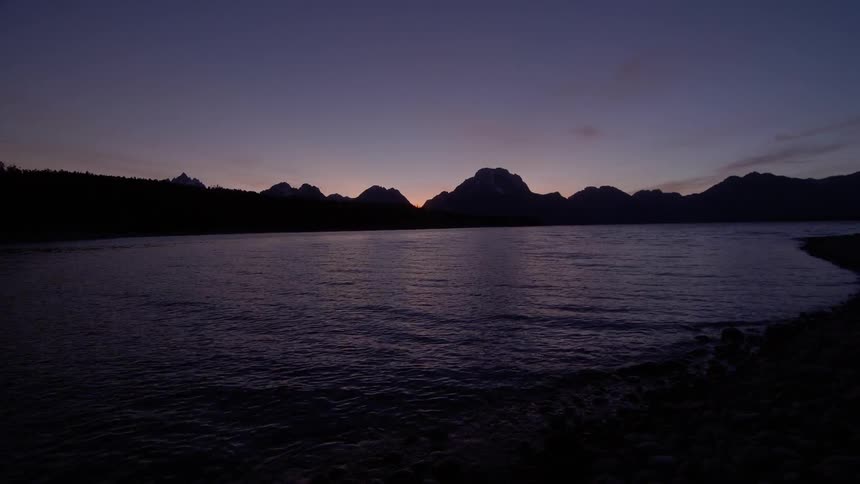 A calm lake hits the shore at night with mountain silhouettes on the horizon