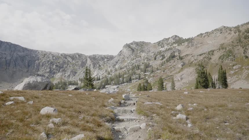 A stone path through a rock strewn grassland at the base of a gray mountain ridge