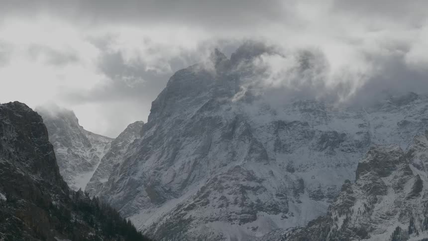 A towering mountain is coated in a solid layer of snow as a layer of clouds passes along the top of it