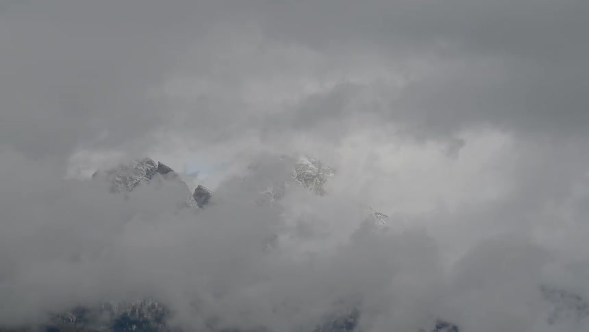 Thick clouds briskly pass in front of a towering snow covered mountain range on a windy day