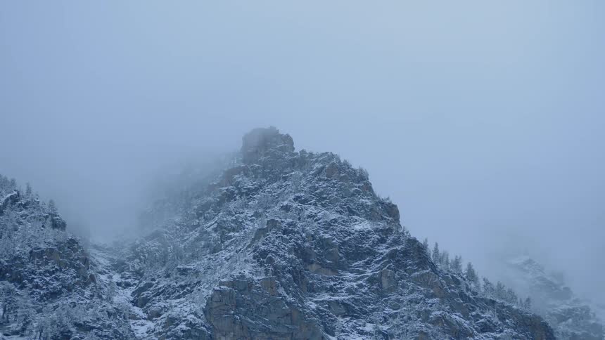 A rocky mountain sparsely littered with trees is covered in ice and snow on a foggy day