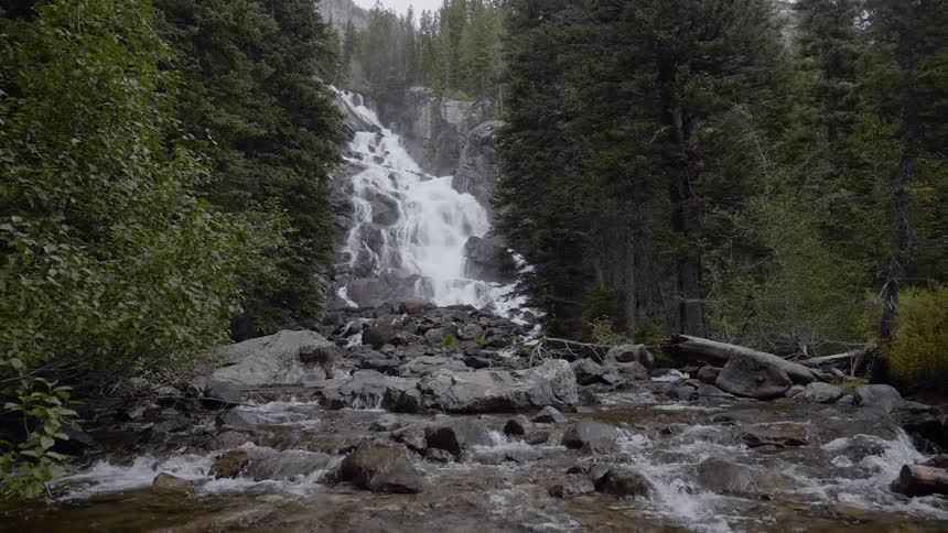 A waterfall cascades down a series of rocks and feeds into a shallow river that runs through a forest in the mountains