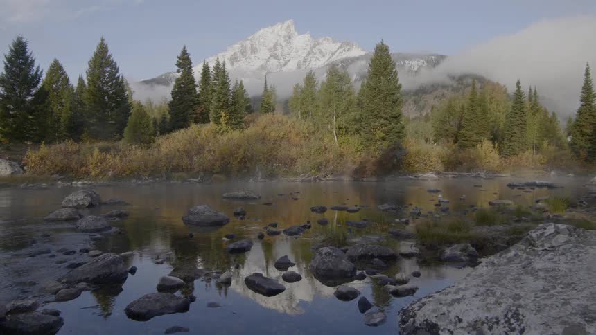 Small and large rocks sits in a shallow creek by a forest and mountain