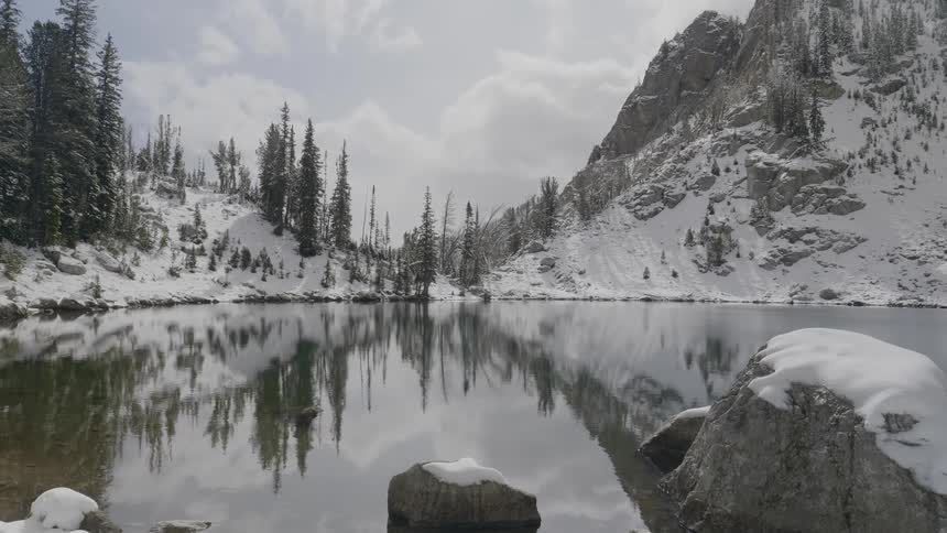 A serene lake is at the base of a snow covered mountain range that has a handful of pine trees lining it