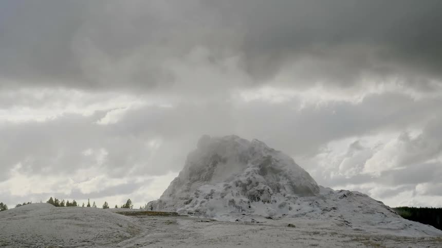 Steam rises from a snow dusted geyser and rocky terrain on a gray cloudy day