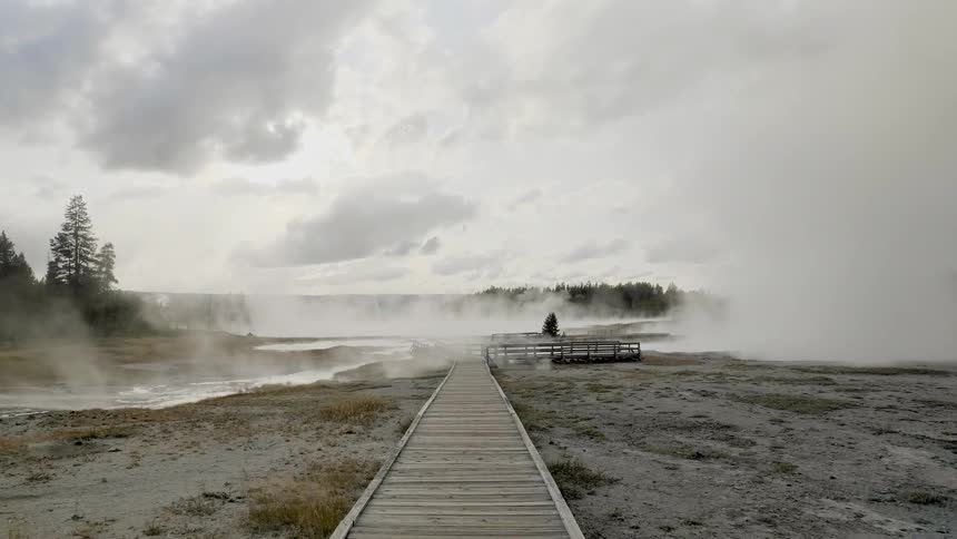 A long wooden walkway leads to a wooden observation deck that is situated near a steaming natural hot spring