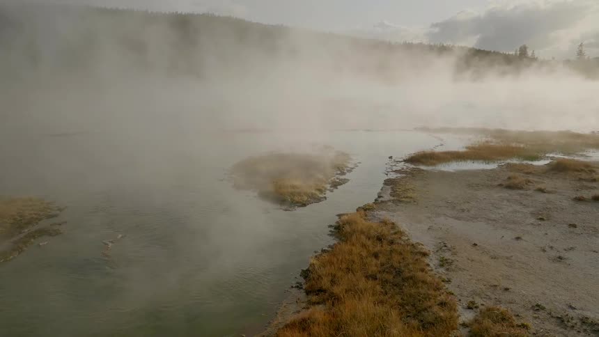 Hot steam hovers of the still waters of a natural hot spring which is surrounded my grassy and muddy shores