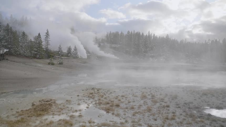 Several massive plumes of steam are billowing from the ground in the distance behind some shallow hot springs
