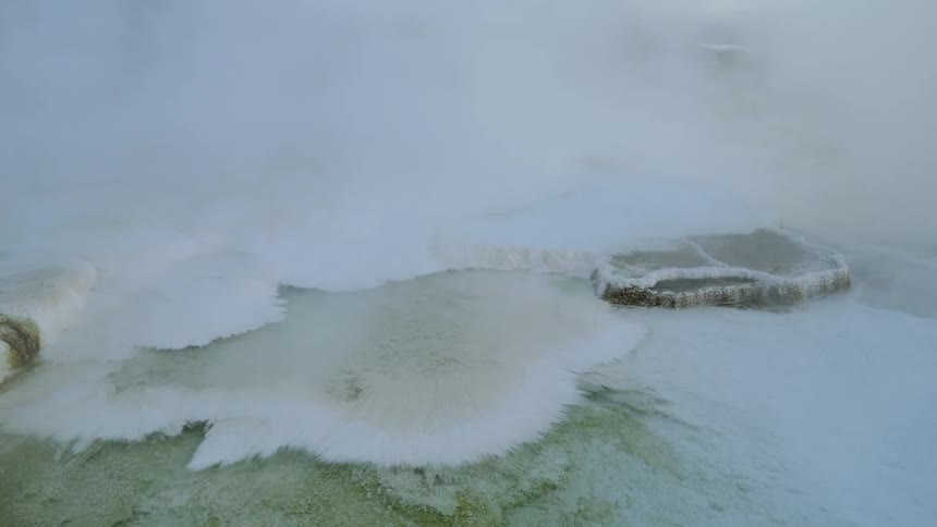 Steam continuously rises from the surface of a natural hot spring that is full of crystallized mineral deposits