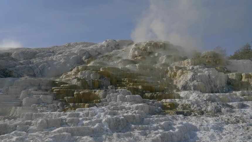 Steam rises from a number of different spots along the sides of a geyser that is covered in layers of mineral deposits