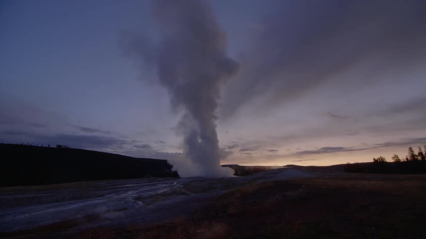 A tall plume of steam rises high into the sky at dusk as it is expelled from a powerful geyser in the ground