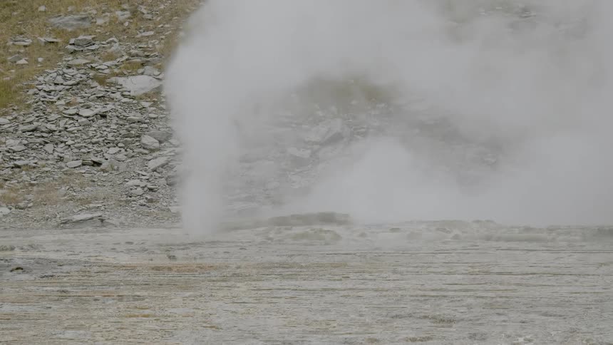 Steam spews from a vent hole in the ground in front of a rocky slope