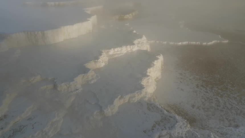 From high above several snow and ice covered cliffs can be seen through a thin layer of clouds passing by