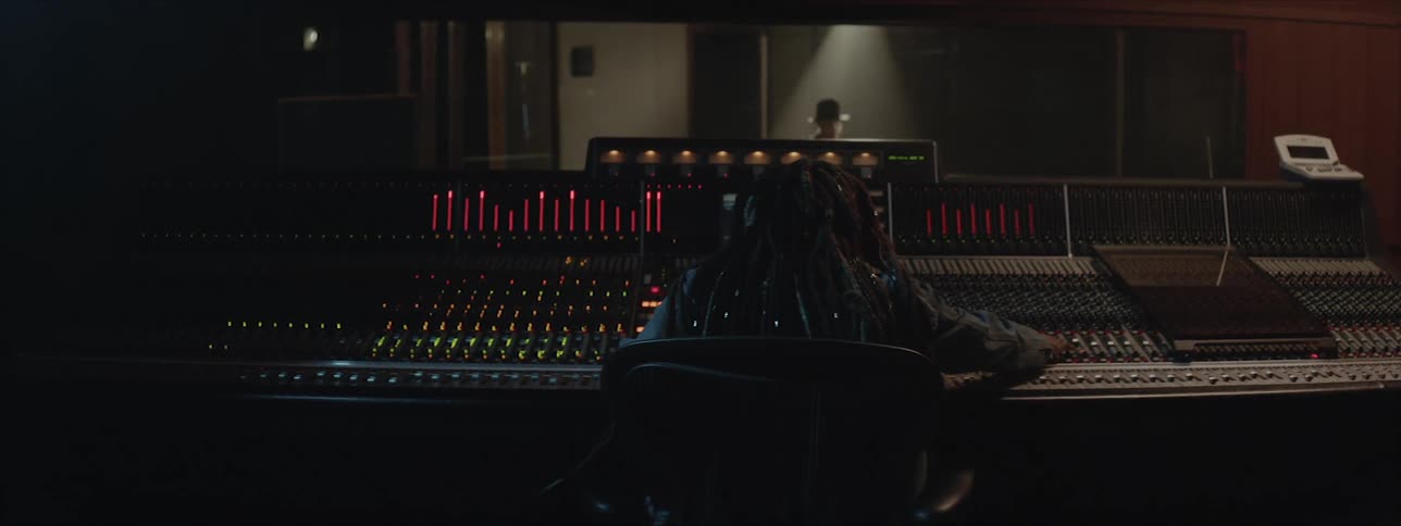 A person with dreadlocks sits at a mixing desk as a drummer wearing a hat sits and plays drums in a studio booth