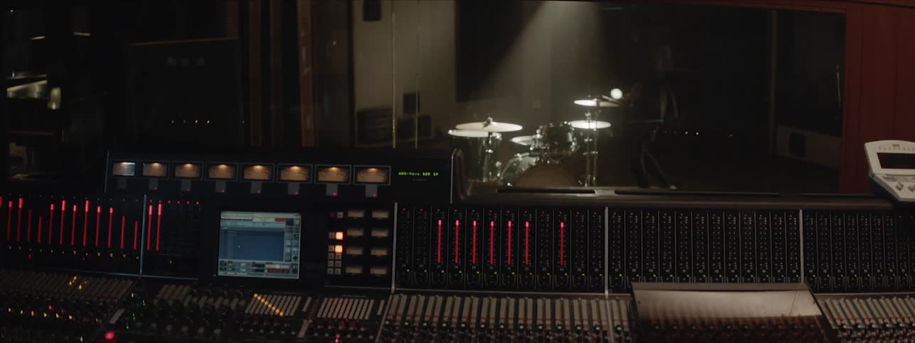 A sound engineer sits in the booth while a man gets on the drums